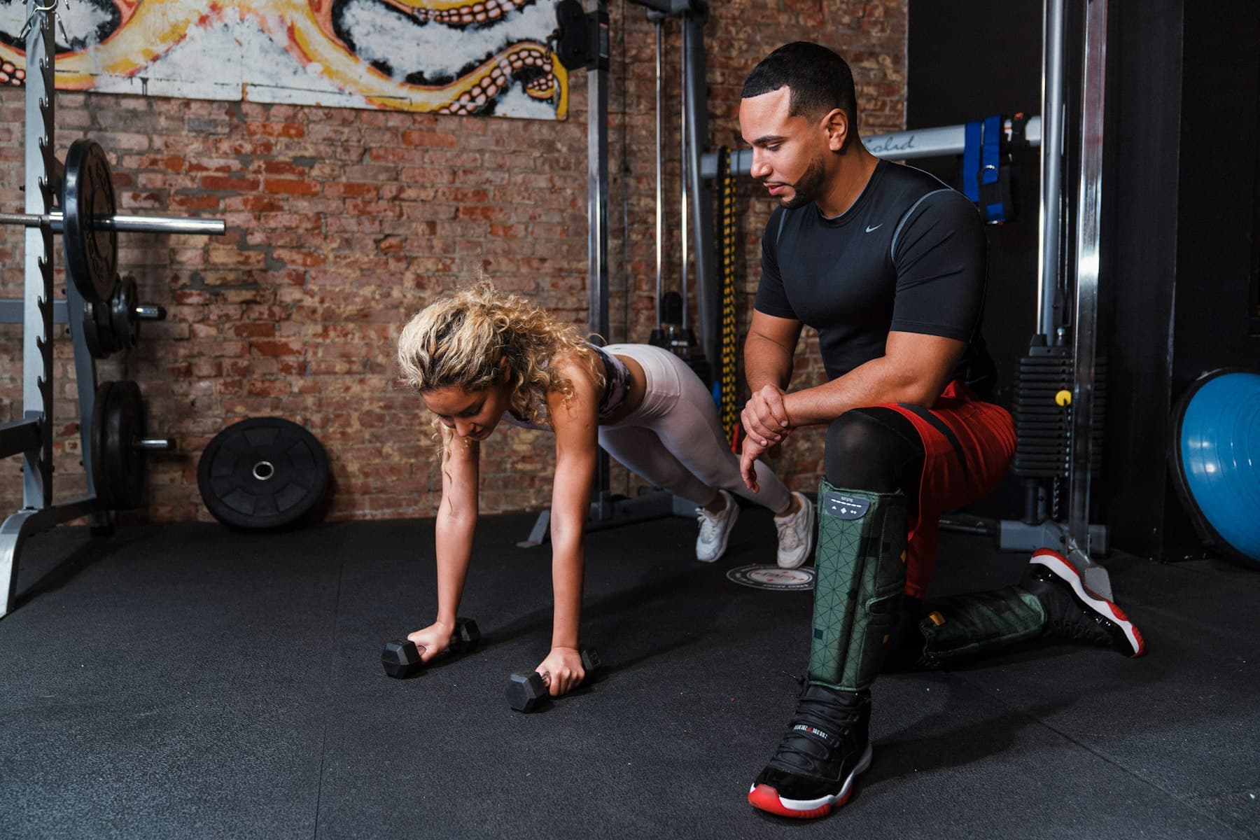 A personal trainer kneels beside his client, watching her form as she holds a plank on dumbbells